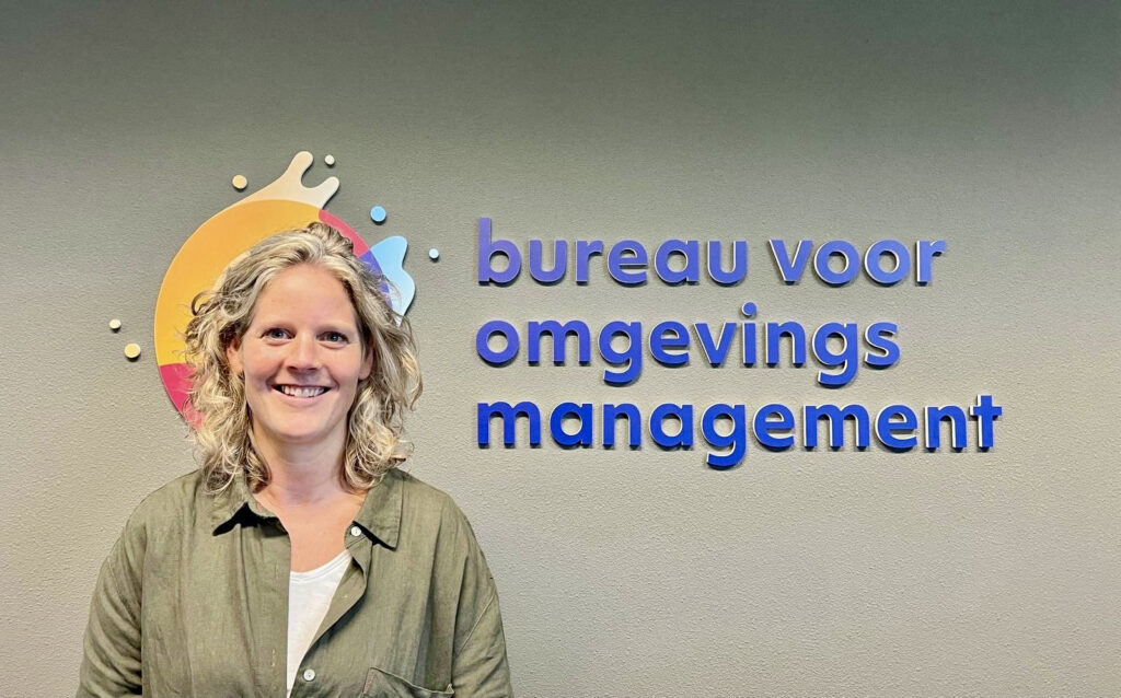 Foto van blonde vrouw die staat voor het logo van bureau voor omgevingsmanagement wat met blauwe letters op de muur geplakt is