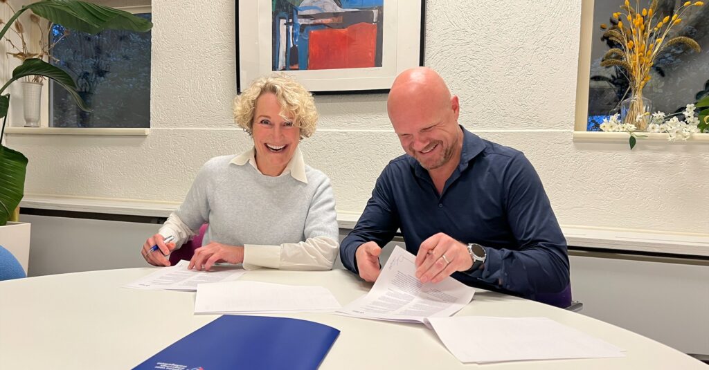 Twee mensen zitten samen aan een tafel terwijl ze documenten ondertekenen; ze lachen ontspannen, met papieren en een blauwe map voor zich, in een lichte ruimte met planten en kunst aan de muur.